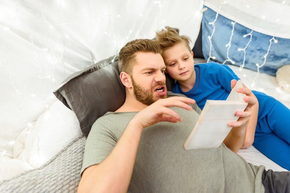 Emotional father and smiling son reading book while lying in blanket fort
